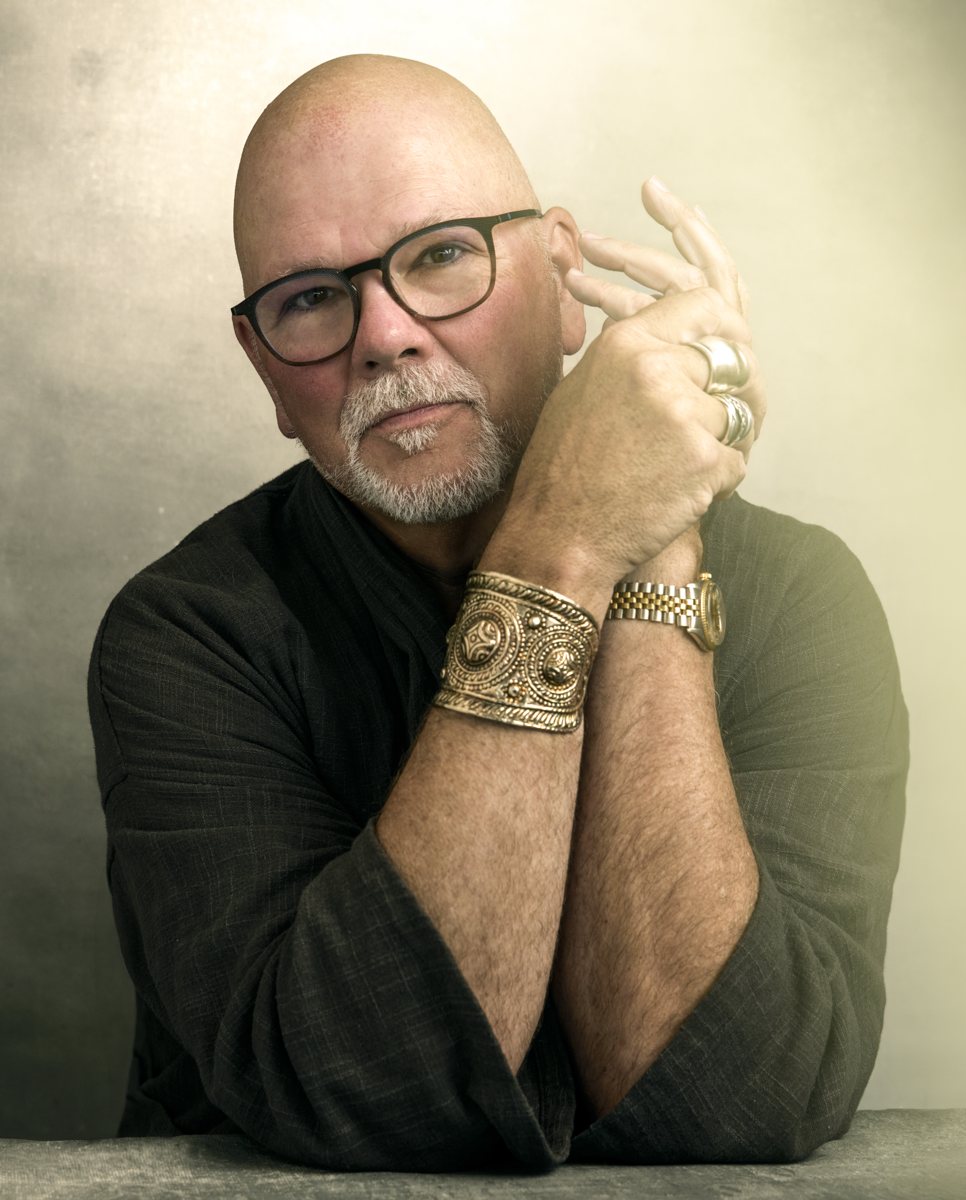 Gary Waddell, seated with hands clasped near his face, wearing a gold cuff, against a warm cream background